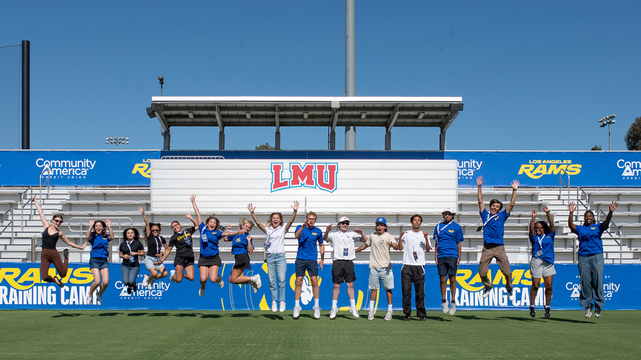 A group of approximately 15 people jump in the air on a grassy athletic field in front of stadium bleachers