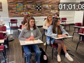 Students sit in desks in a classroom. Numbers overlay, showing this is a shot from a film in development.