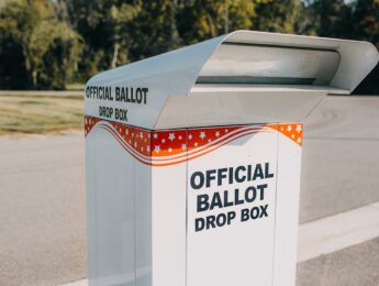 A labeled official ballot drop box stands in an open area with trees in the background. The drop box is white with red and blue stars and stripes.