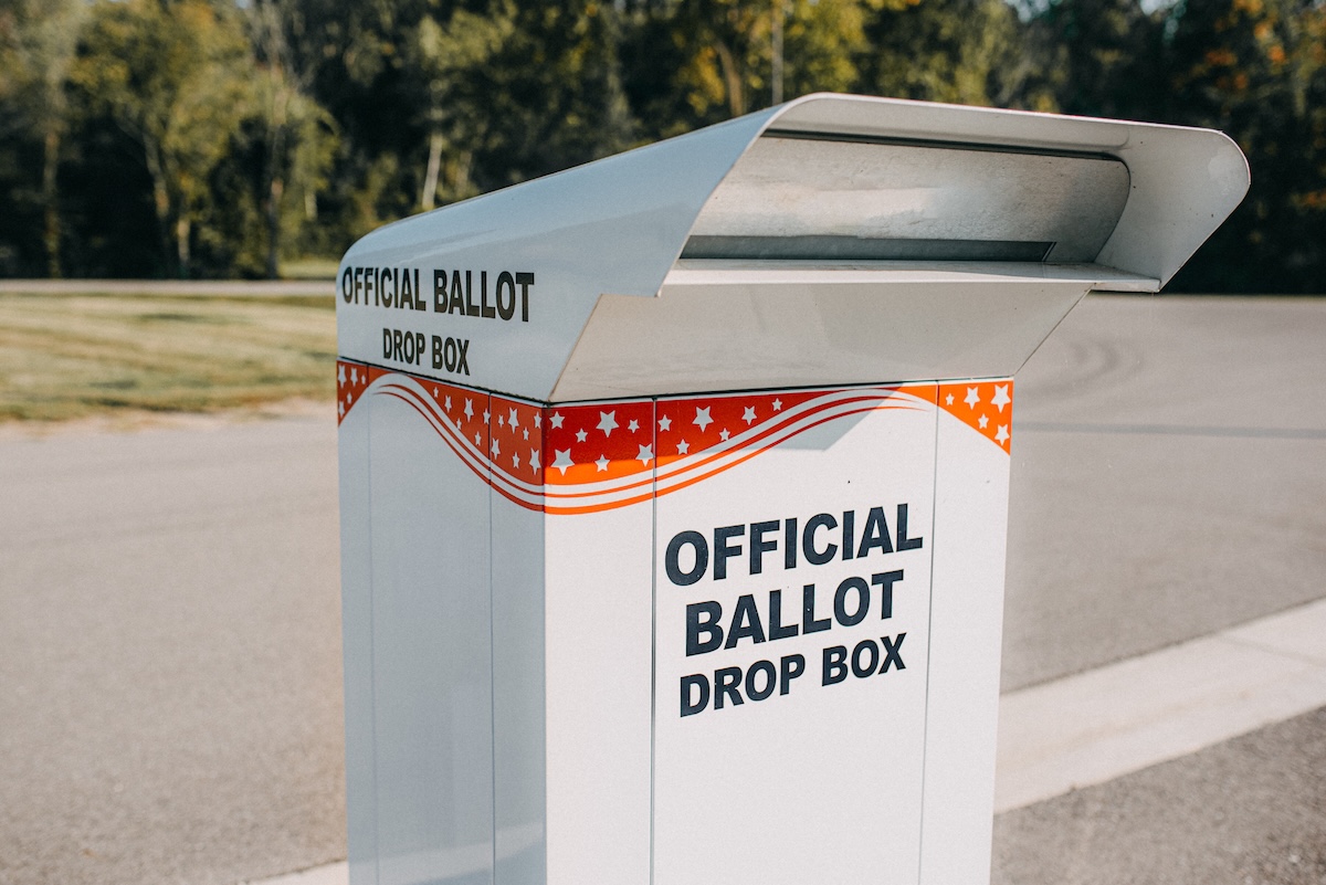 A labeled official ballot drop box stands in an open area with trees in the background. The drop box is white with red and blue stars and stripes.
