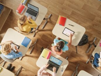 Top View Shot in Elementary School Computer Science Classroom: Children Sitting at their School Desk Using Personal Computers and Digital Tablets for Assignments.