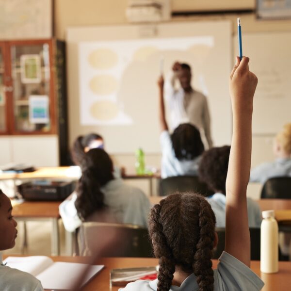 Rear view of girl raising hand and answering while sitting next to friend during lecture in classroom