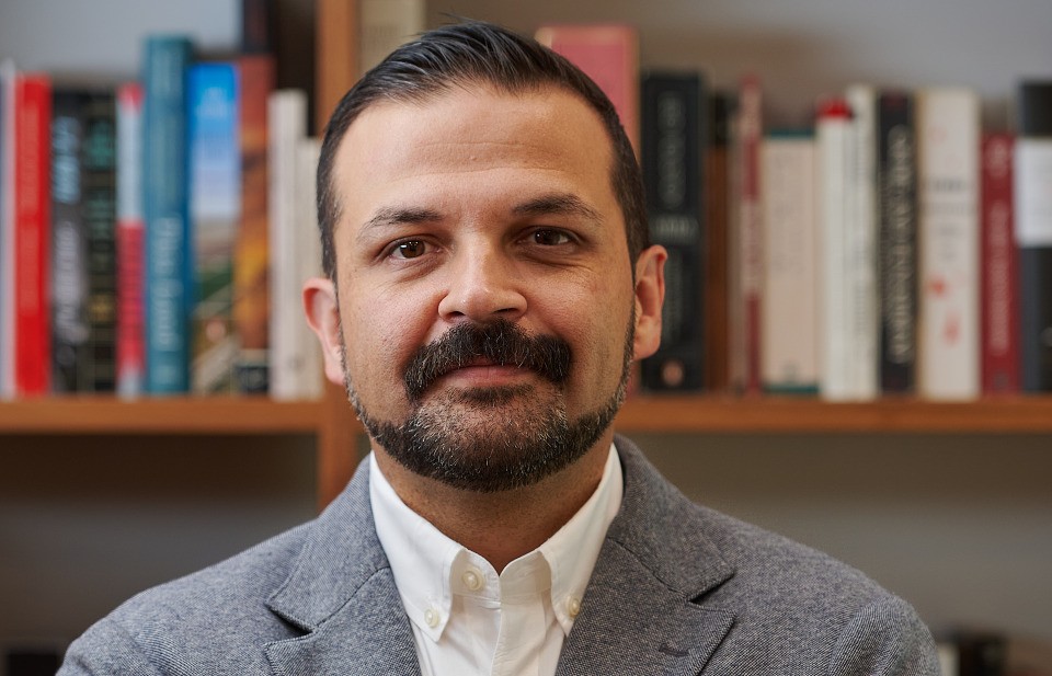 Man with dark hair, wearing gray blazer and white button up pictured in front of bookshelves