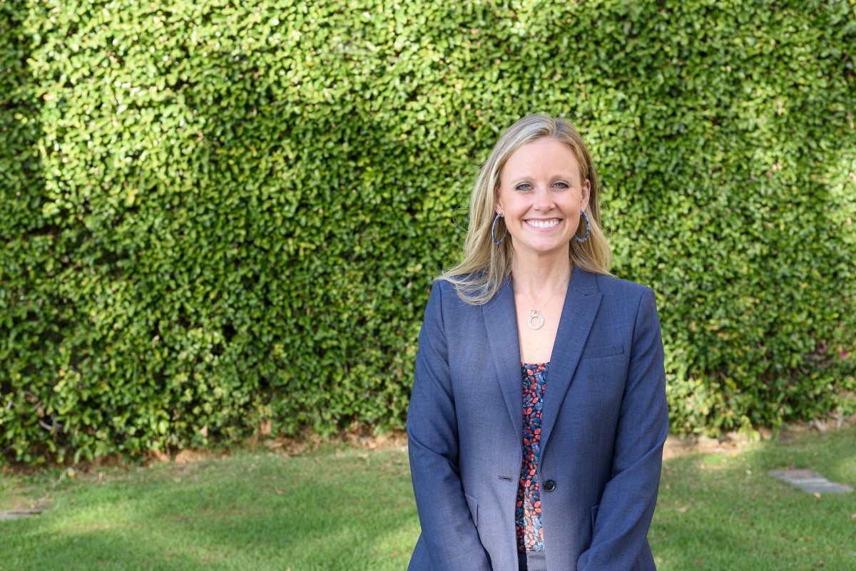 Brianne Gilbert wearing navy blazer, smiling, against green hedge.