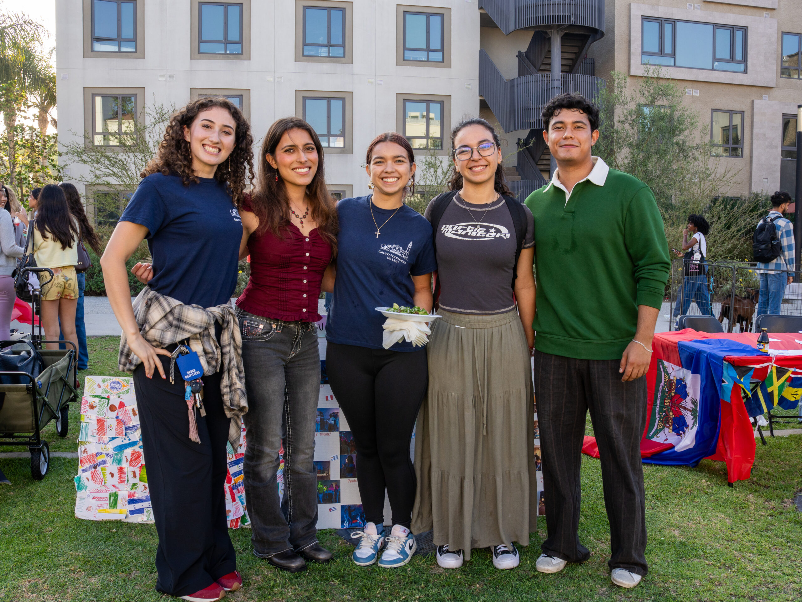 Five young adults smile for a group photo at an outdoor cultural celebration. They stand on a grassy lawn in front of a modern apartment building, with colorful flags, decorated tables, and artwork displayed behind them.