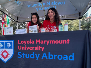 Two people sit behind a Loyola Marymount University Study Abroad information table under a canopy tent at an outdoor campus event.