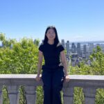 A woman in a black tee and pants standing on a balcony with a cityscape behind her.