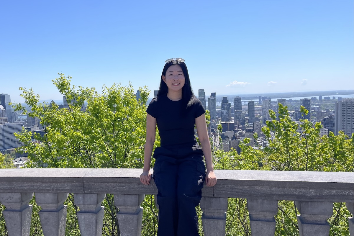 A woman in a black tee and pants standing on a balcony with a cityscape behind her.