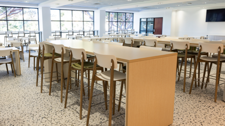 Bright, newly renovated dining hall featuring a mix of communal high-top tables and standard seating. Large windows line the space, bringing in abundant natural light and views of trees outside. The interior includes light wood furnishings, white chairs, terrazzo-style flooring, and a clean, contemporary design.
