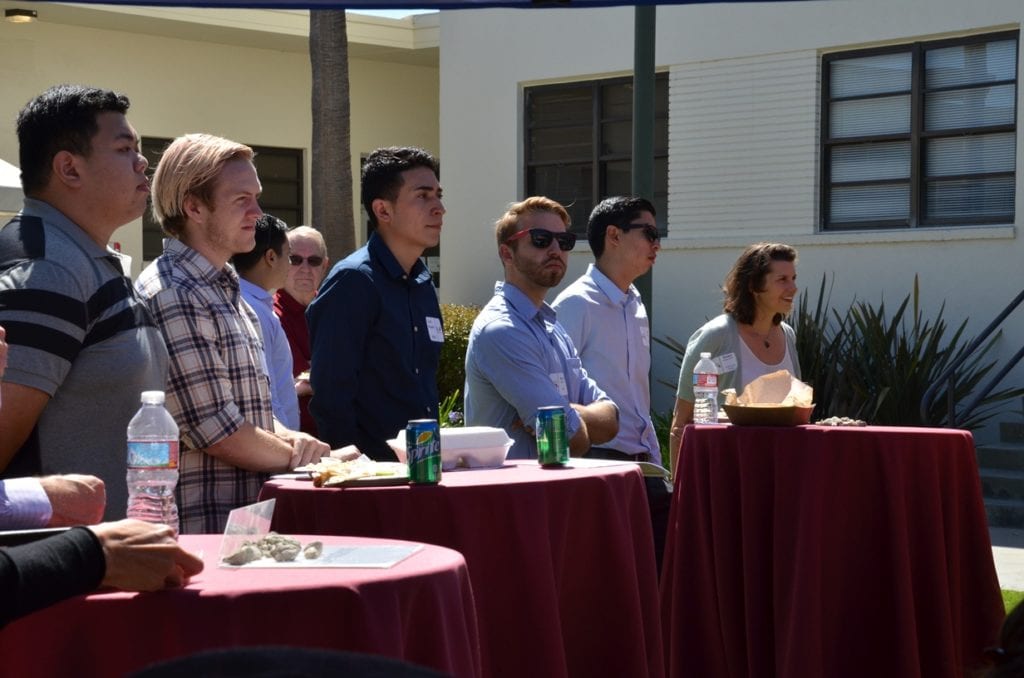 Students standing behind tables and listening intently to an alumnus speak