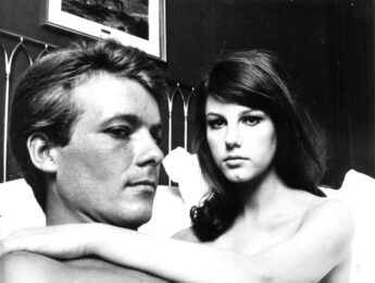 A black and white film still of a man and woman laying in bed with the woman looking into the camera