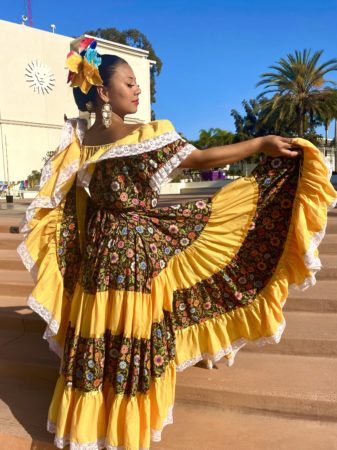 A student stands outside of Malone Student Center in a yellow dress to perform with Grupo  de Folklórico.