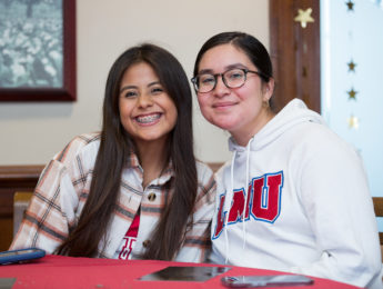 Two students sit inside the Hill on LMU's campus enjoying an event while wearing LMU gear.