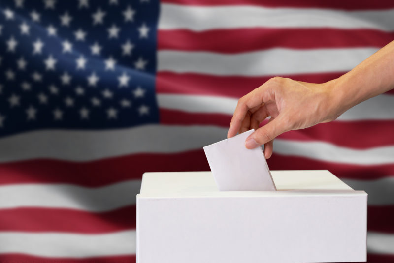 Close-up of man casting and inserting a vote and choosing and making a decision what he wants in polling box with United States flag blended in background