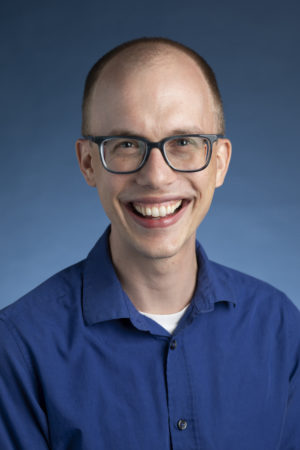 A headshot of Timothy Williamson in a blue shirt against a blue background