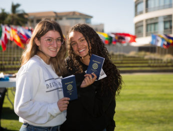 Two female students holding a passport stand in front of LMU's library and international flags.