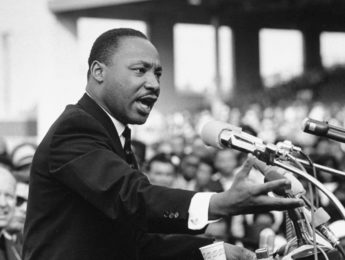 Dr. Martin Luther King, Jr. speaks at a podium outside to a crowd.