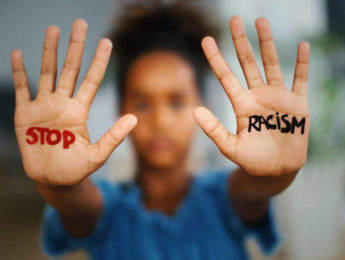 African american girl standing indoors and looking at the camera. She's showing an message that says Stop Racism on her hands.