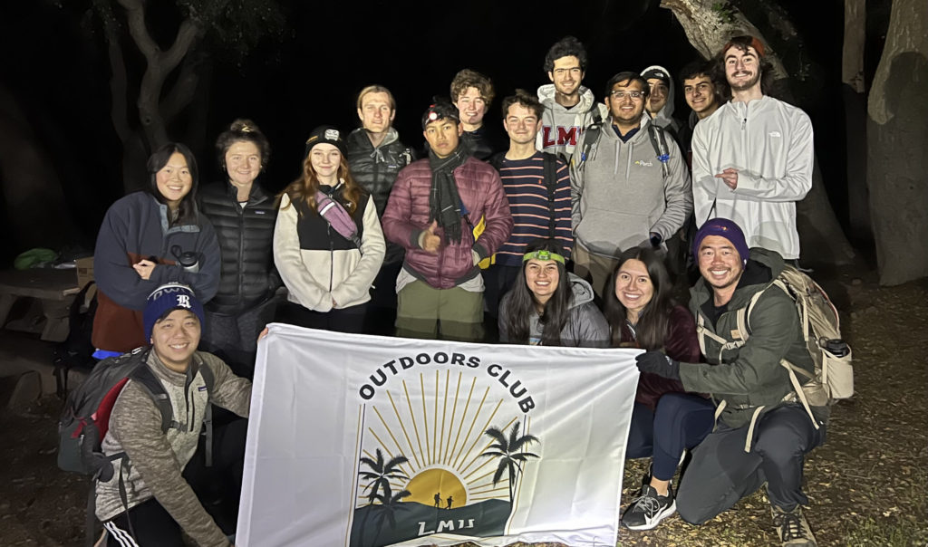 Students and staff from the LMU Outdoor Club stand outside with an Outdoor Club flag after a nighttime hike.