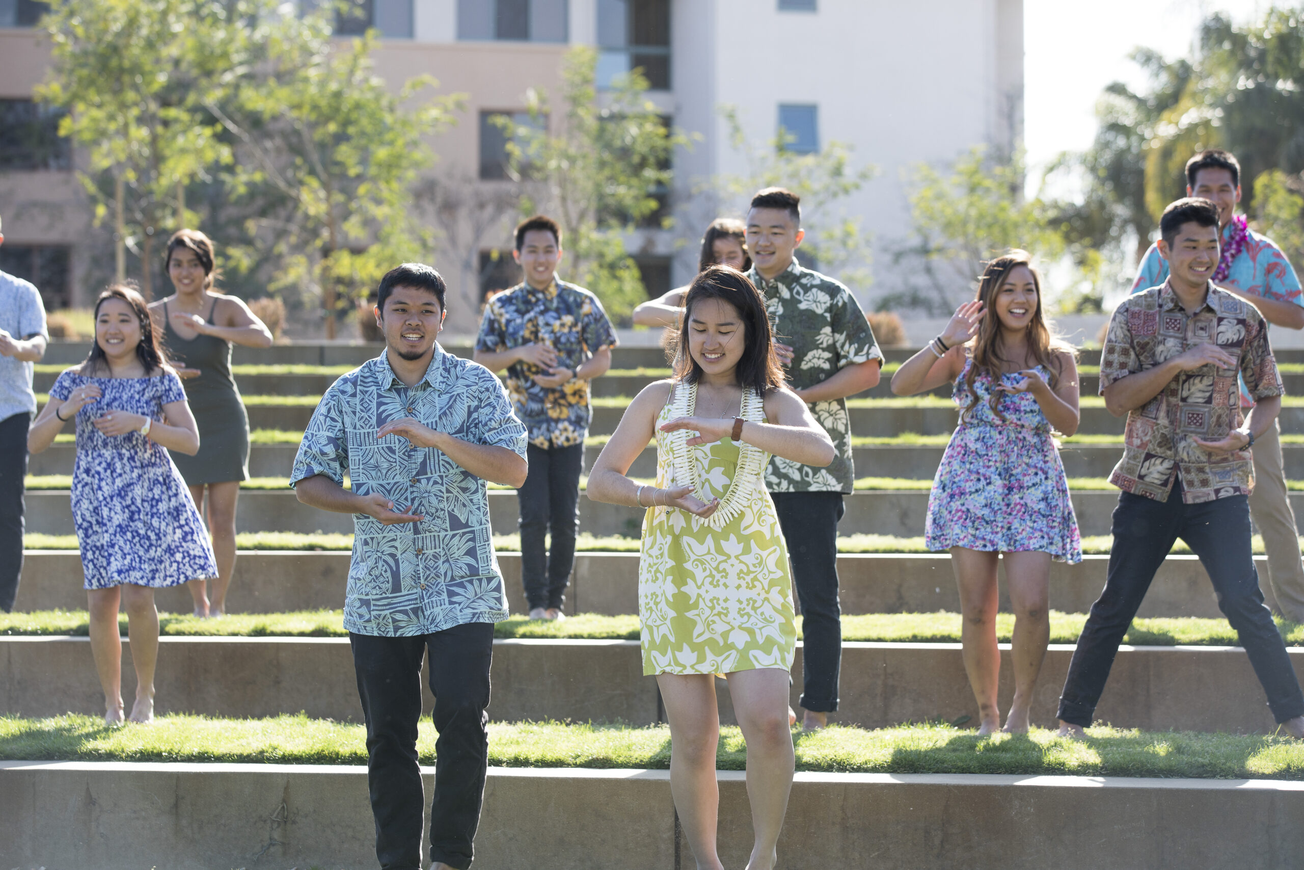 Students stand on green grassy steps wearing blue and yellow Hawaiian style clothes dancing Hula.