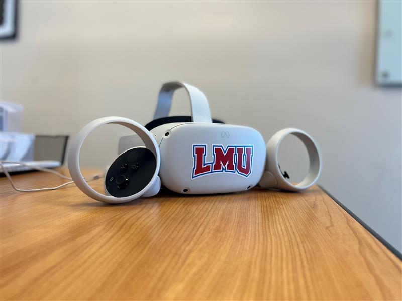 A virtual reality white headset with an LMU logo and controllers sit on a wood grain table.