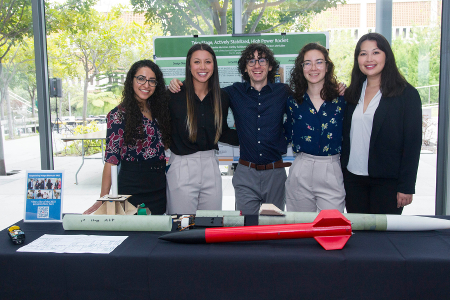 Five students smile next to their capstone project titled 'Two Stage, Actively Stabilized, High Power Rocket'. The rocket is on the table in front of them.