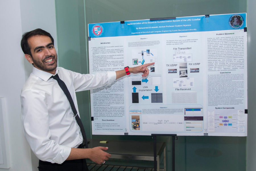 A student smiles standing next to the poster board for their project titled 'Implementation of the Downlink Communication System of the LMU Cubesat'
