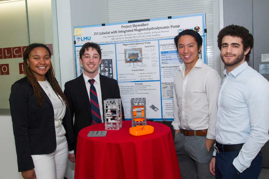 Four students smile standing next to the poster board for their project titled 'Project Skywalker: 2U CubeSat with Integrated Magnetohydrodynamic Pump'. The prototype for their project his on the table in front of them.