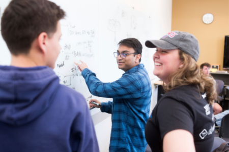 A group of hackathon participants smile next as one writes on a whiteboard