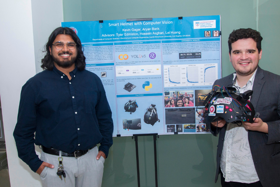 Two students smile as they stand next to the poster board for their project titled 'Smart CV Helmet', one of whom is holding the helmet.