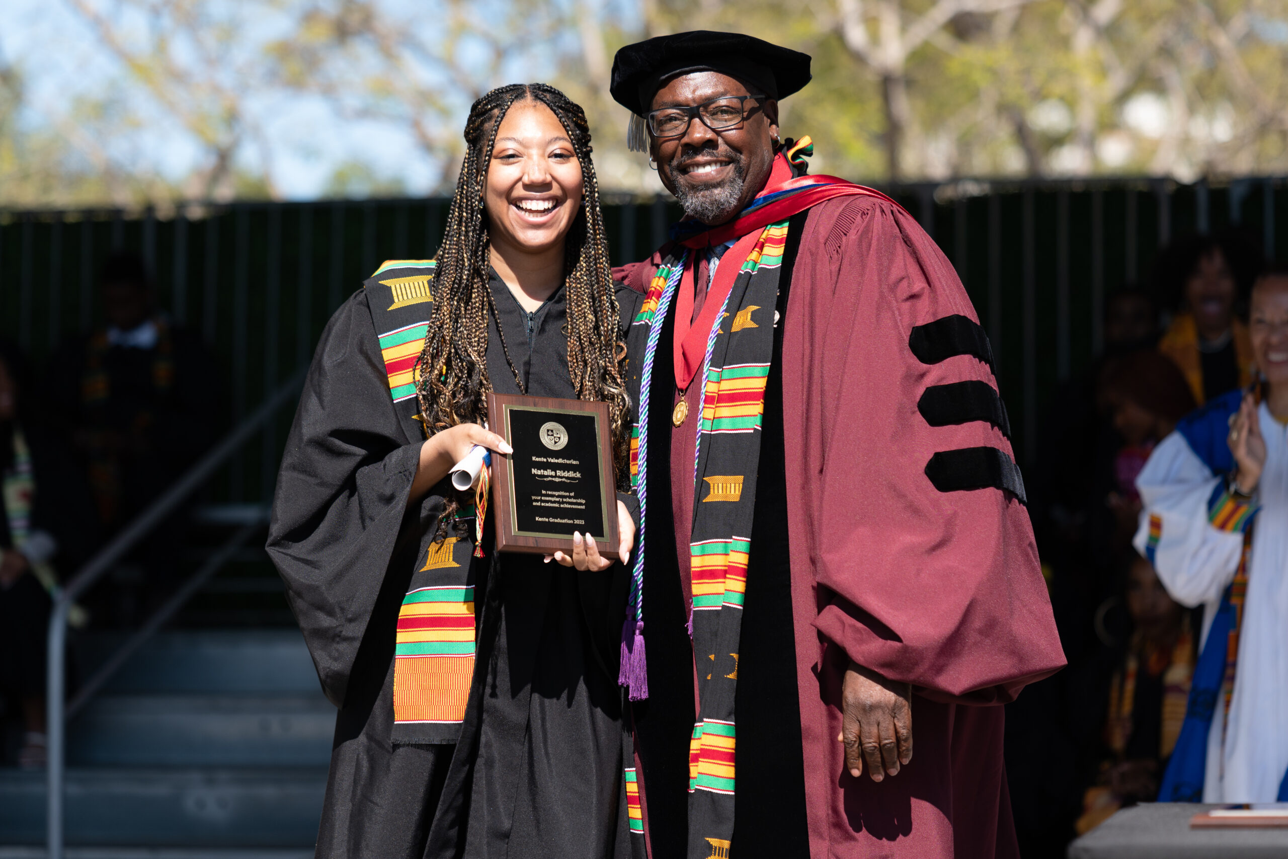 A student and dean stand to pose for a photo of their award on a stage outside.