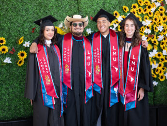Students in graduation regalia stand in front of the a yellow floral and green backdrop.