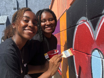 Two students pose in front of a mural and paint the wall to refresh it during an annual day of service.