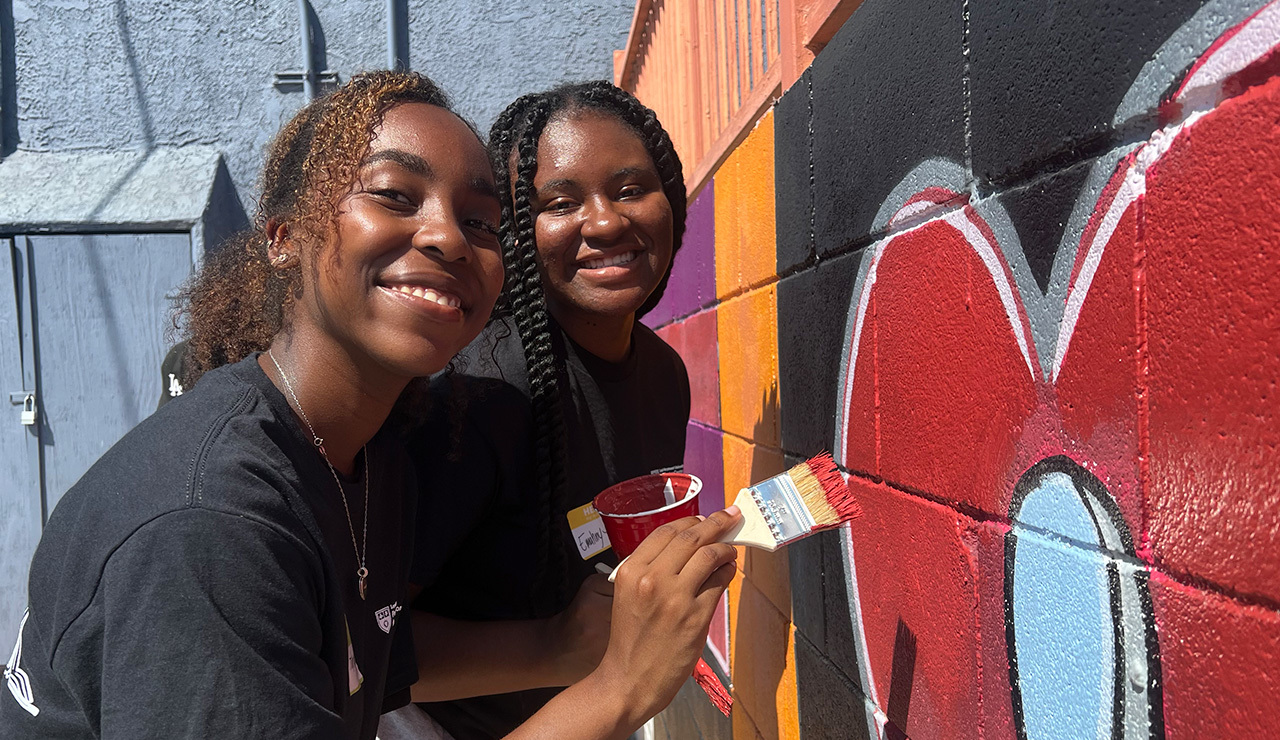 Two students pose in front of a mural and paint the wall to refresh it during an annual day of service.