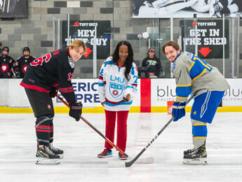 One hockey player in a black and red jersey and one in a blue and grey stand with an official dressed to drop the puck for the start of the game.