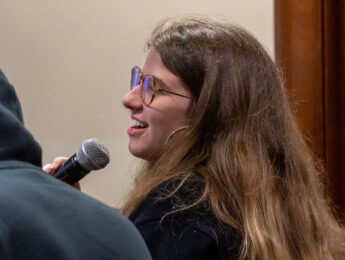A student with long brown hair and glasses holds a microphone to speak at an event.