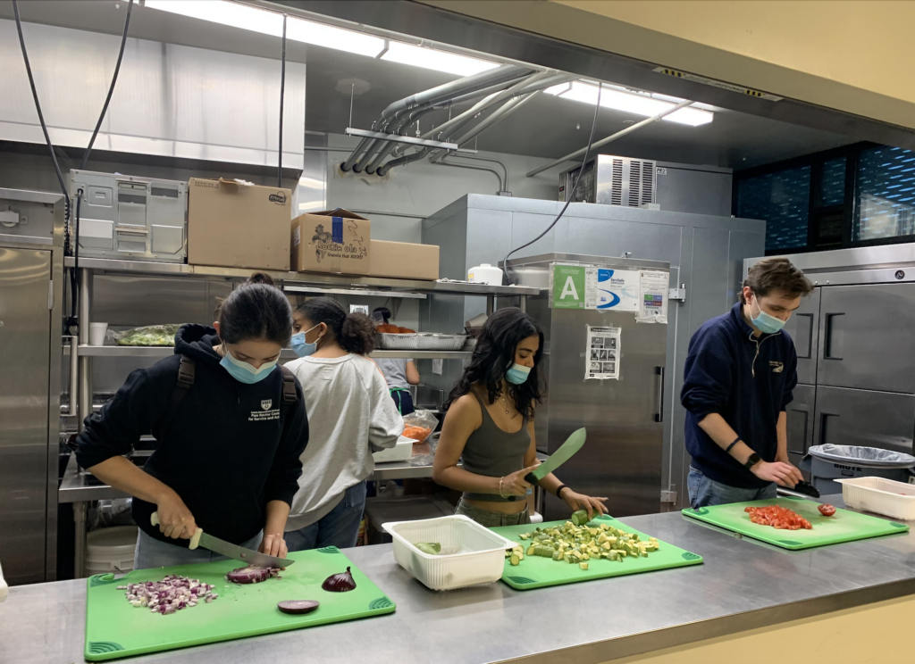 Four students are standing in a kitchen using a knife to cut up vegetables and prepare food.