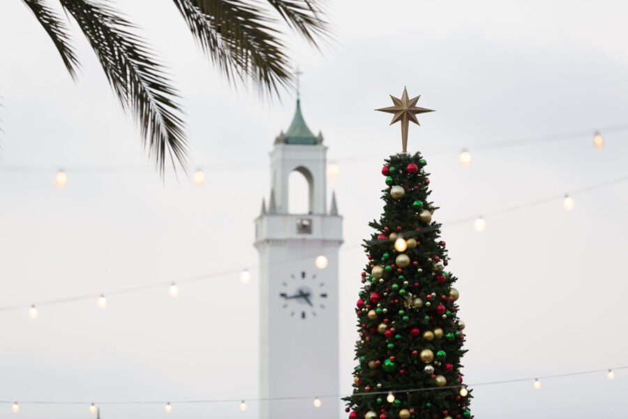 A green Christmas tree with star topper is in front of the clock tower outside.