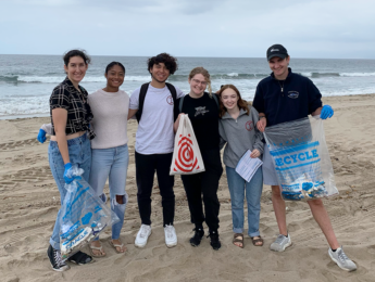 Students stand holding bags on the sand of the beach during a beach clean-up.