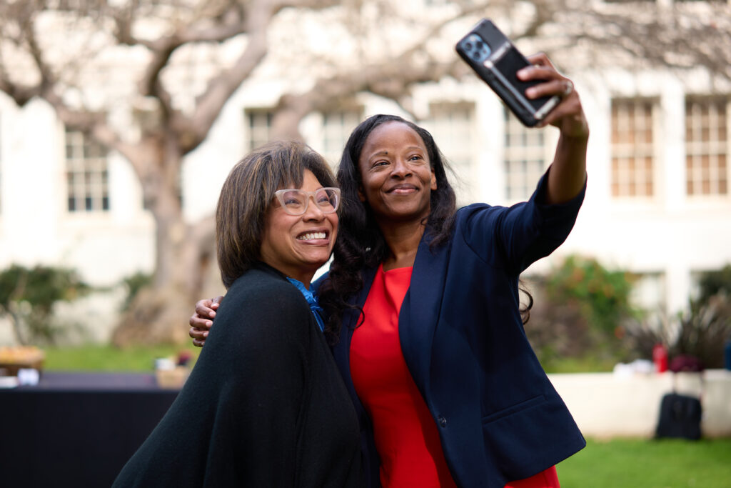 Two women in black and a dark blue jacket with red dress pose for a selfie outside.