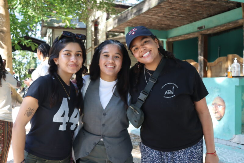 Three female students in black shirts and a grey shirt and vest pose outside.
