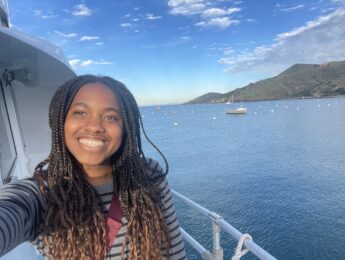 A student takes a selfie on a ferry boat with the blue water and Catalina Island in the background.
