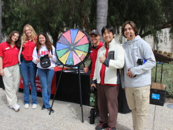 Students pose outside with a colorful prize wheel.