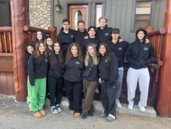 Students in black sweatshirts pose outside of a brown wooden home.