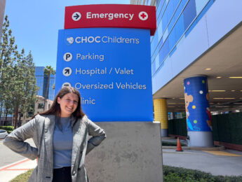 A student in a grey suit stands in front of a blue and red sign for Children’s Hospital of Orange County.