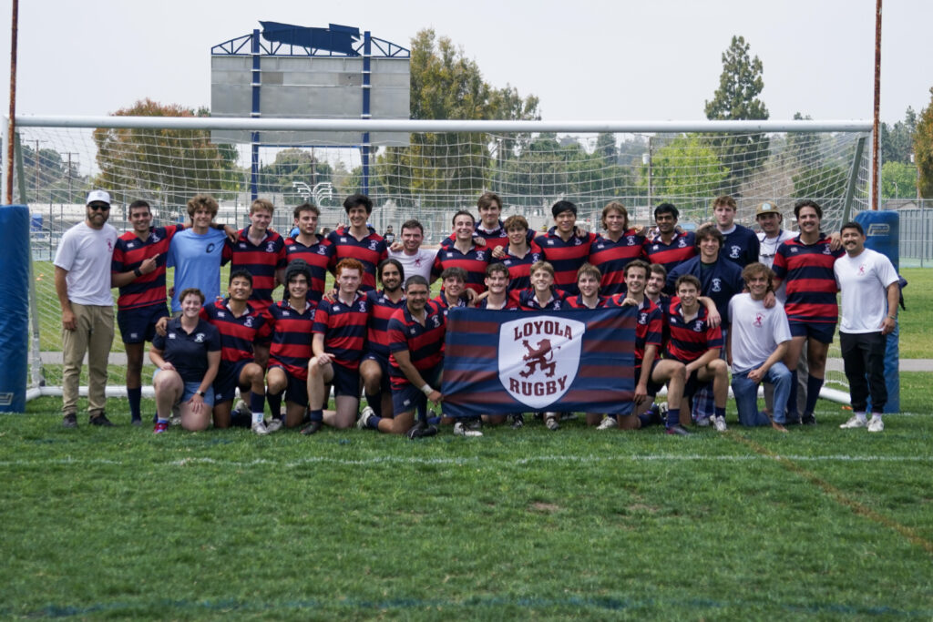 Students and coaching staff pose on a green field holding a blue and red flag.