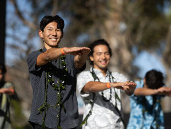Male students in Hawaiian shirts dance with their arms out to perform hula outside.